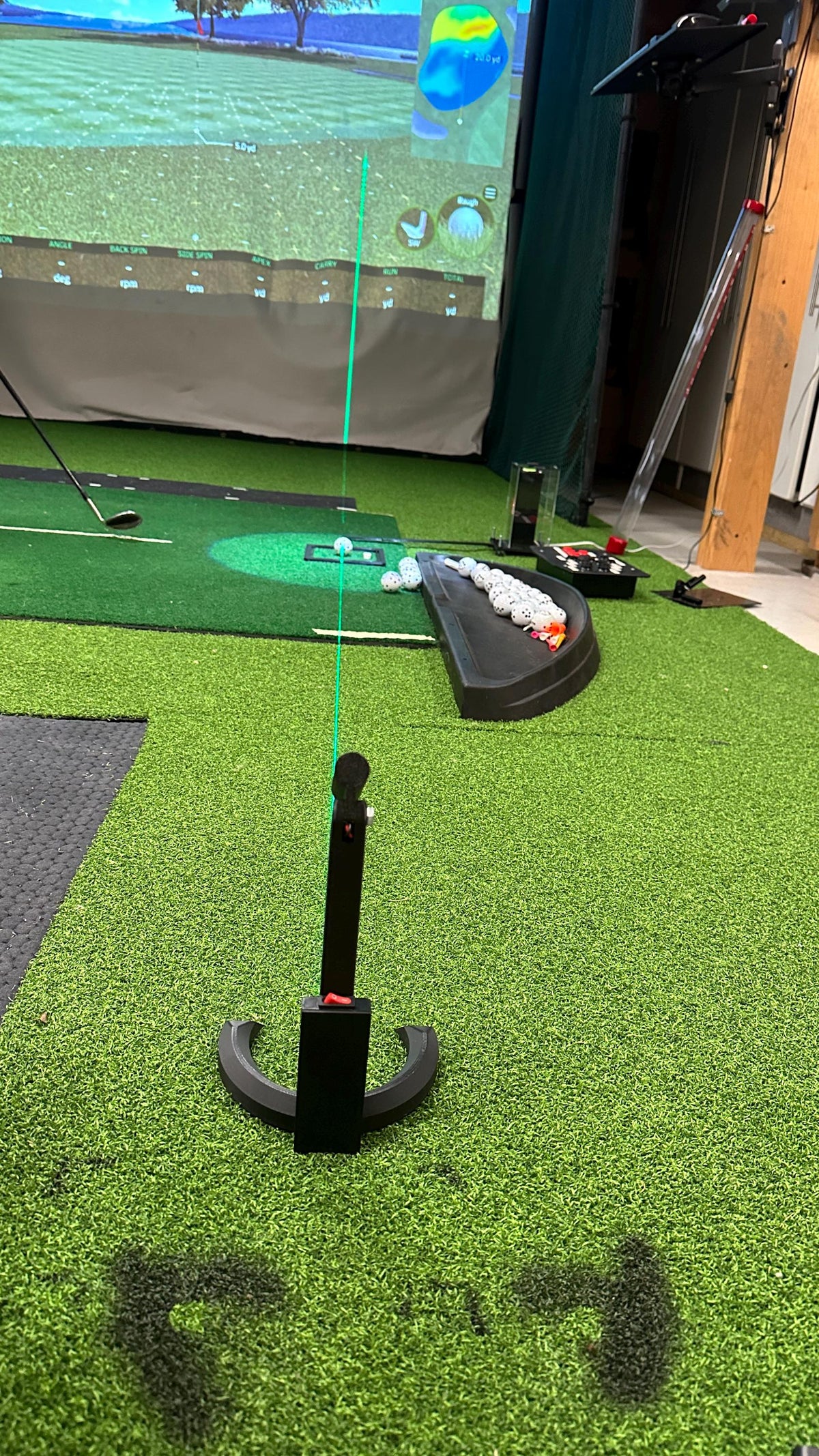 Laser alignment tool positioned on a golf mat in front of a simulator screen